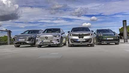 Vier Elektro-SUVs – Audi Q6 SUV e-tron quattro, BMW iX3 50 xDrive, Mercedes GLC 400 4matic EQ und Porsche Macan 4S – nebeneinander auf einer offenen Fläche, fotografiert aus niedriger Perspektive, mit Himmel und Wolken im Hintergrund.