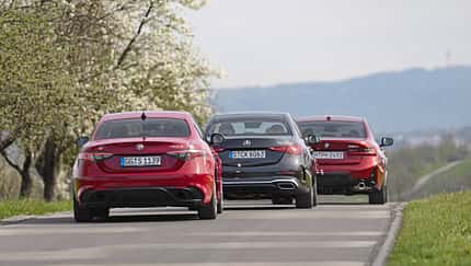 Drei Limousinen, Alfa Romeo Giulia 2.2 Diesel Q4, BMW 320d und Mercedes C 220d, fahren auf einer Landstraße. Die Fahrzeuge sind von hinten zu sehen, umgeben von blühenden Bäumen und einer hügeligen Landschaft.