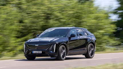 Cadillac Lyriq-V fährt auf einer Landstraße, Seitenansicht, schwarze Lackierung, grüne Vegetation im Hintergrund.
