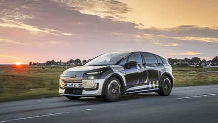 Audi A2 e-tron, Erlkönig, Elektroauto, Audi, Kompaktwagen, Tarnfahrzeug, Prototyp, Elektromobilität, Straßenfahrt, Sonnenuntergang Audi A2 e-tron Erlkönig (Tarnfahrzeug) fährt auf Landstraße bei Sonnenuntergang. Das Fahrzeug trägt eine auffällige Tarnfolierung, im Hintergrund ist eine ländliche Szenerie mit Häusern und Wiesen zu sehen.