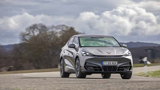 Der Cupra Tavascan Endurance fährt auf einer Landstraße in ländlicher Umgebung. Das Fahrzeug ist frontal leicht von der Seite zu sehen, die Umgebung zeigt Bäume und einen bewölkten Himmel.
