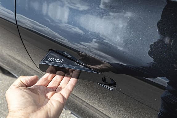 Eine Hand greift nach einem Türgriff eines Autos, auf dem das Logo 'smart' zu sehen ist. Die Autotür spiegelt den Himmel und die Person wider.