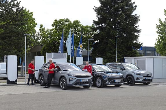 Drei Elektroautos werden an einer Ladestation von drei Personen in roten Hemden geladen. Im Hintergrund sind Bäume, Fahnen und ein Gebäude zu sehen.
