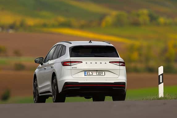 Das Bild zeigt einen weißen Skoda Enyaq RS 79 kWh von hinten auf einer Landstraße in ländlicher Umgebung. Im Hintergrund sind Felder und sanfte Hügel zu sehen.