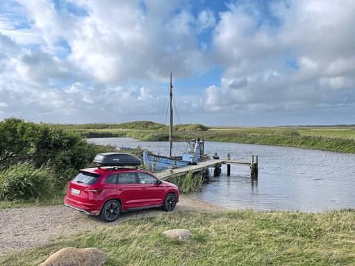 Skoda Karoq 2.0 TDI mit Dachbox an einem kleinen Bootsanleger in Dänemark, im Vordergrund ein rotes SUV, im Hintergrund ein Boot am Steg und weite Wiesenlandschaft unter bewölktem Himmel.