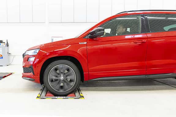 Rotes Skoda Karoq SUV, Seitenansicht, auf Rollenprüfstand in heller Werkstattumgebung, Fahrer am Steuer.