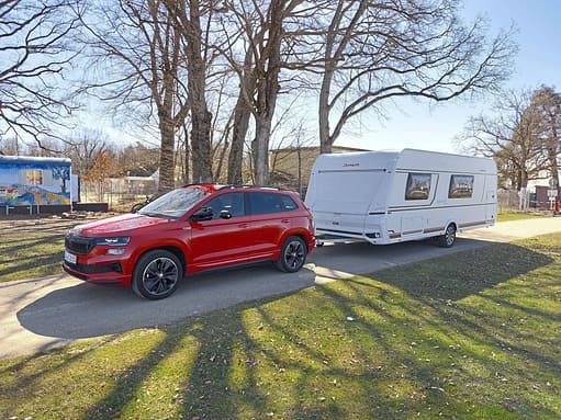 Ein roter Skoda Karoq zieht einen weißen Dethleffs Camper Wohnwagen auf einem Campingplatz. Im Hintergrund sind Bäume und ein bunt bemalter Bauwagen zu sehen. Die Aufnahme entstand bei Tageslicht im Frühjahr.