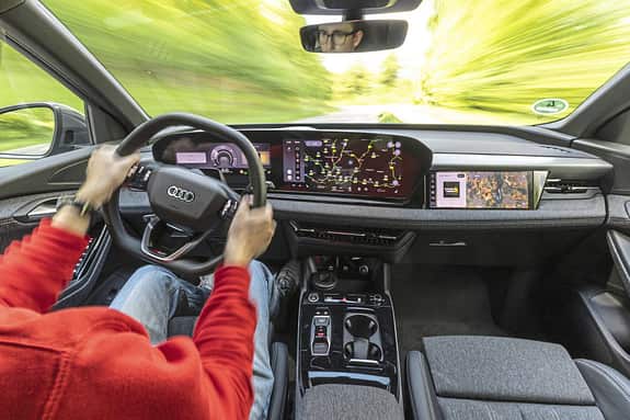 Innenraum und Cockpit des Audi Q6 SUV e-tron quattro während der Fahrt, Fokus auf digitales Armaturenbrett und Fahrer.