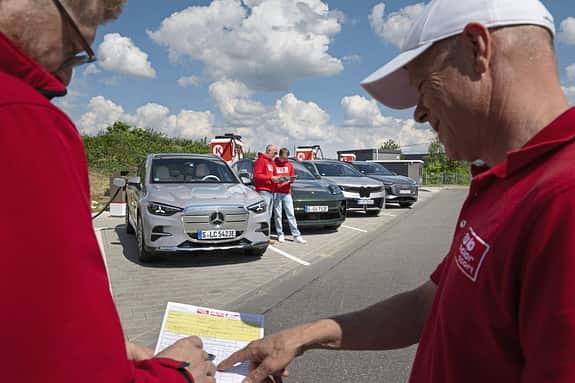 Vergleichstest von vier Elektro-SUVs (BMW i5 30xDrive, Mercedes GLC 400 4MATIC EQ, Audi Q6 SUV e-tron quattro, Porsche Macan 4S) an einer Ladestation mit mehreren Personen im Vordergrund.