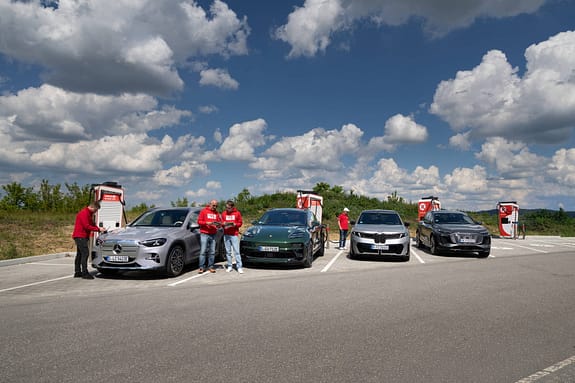 Vier Elektro-SUVs – BMW i5 30xDrive, Mercedes GLC 400 4MATIC EQ, Audi Q6 SUV e-tron quattro und Porsche Macan 4S – stehen nebeneinander an Ladesäulen auf einem Parkplatz, begleitet von mehreren Personen in roten Oberteilen, die Messungen oder Notizen durchführen. Der Himmel ist leicht bewölkt.