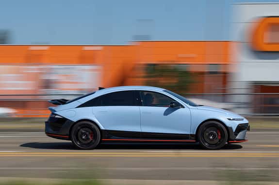 Hyundai Ioniq 6N AWD fährt auf einer Straße vor einem orangefarbenen Gebäude in der Stadt. Das sportliche Elektrofahrzeug ist in Seitenansicht zu sehen, dynamisch in Bewegung.