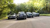 Vier kompakte SUV-Modelle – Dacia Bigster Hybrid, Opel Frontera Hybrid, Hyundai Kona 1.6-GDI HEV und Skoda Karoq 1.5 TSI – fahren nebeneinander auf einer Straße durch eine bewaldete Umgebung.