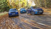 Drei kompakte SUV-Modelle – VW Taigo 1.5 TSI, Opel Mokka Hybrid und Ford Puma 1.0 EcoBoost – fahren nebeneinander auf einer herbstlichen Landstraße.