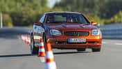 Mercedes Benz SLK 230 Kompressor fährt auf Teststrecke durch Slalom mit Pylonen. Cabrio in orange, Frontansicht, Fahrer sichtbar.