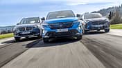 Drei aktuelle Hybrid-SUVs – BMW X1 xDrive 30e, Renault Rafale Hyper Hybrid E-Tech und Cupra Terramar VZ e-Hybrid – fahren nebeneinander auf einer Landstraße. Die Fahrzeuge sind aus der Frontperspektive in Bewegung aufgenommen.