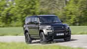 Land Rover Defender 130 D250, SUV, Geländewagen, Allradantrieb, Offroad, Land Rover, Defender, Straßenfahrt, Automobil, Fahrzeug Land Rover Defender 130 D250 fährt auf einer Landstraße, seitliche Frontansicht, grüne Natur im Hintergrund.