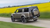 Land Rover Defender 130 D250 fährt auf einer Landstraße an einem blühenden Rapsfeld vorbei.