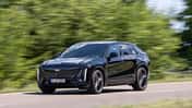 Cadillac Lyriq-V fährt auf einer Landstraße, Seitenansicht, schwarze Lackierung, grüne Vegetation im Hintergrund.