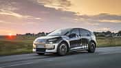 Audi A2 e-tron Erlkönig (Tarnfahrzeug) fährt auf Landstraße bei Sonnenuntergang. Das Fahrzeug trägt eine auffällige Tarnfolierung, im Hintergrund ist eine ländliche Szenerie mit Häusern und Wiesen zu sehen.