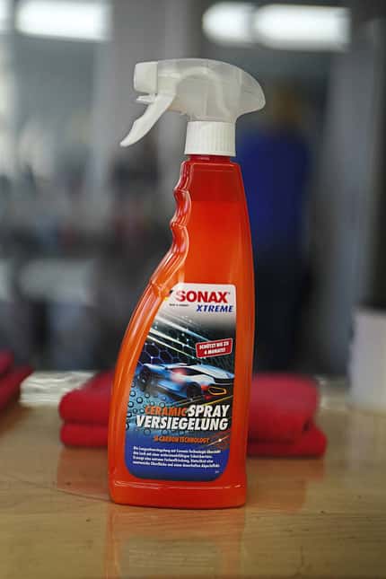 SONAX XTREME Ceramic Spray Versiegelung, Keramikversiegelung, Autopflege, Sprühversiegelung, Fahrzeugpflege, Si-Carbon Technology, Lackschutz, Langzeitversiegelung, Pflegeprodukt, Autozubehör Produktfoto einer SONAX XTREME Ceramic Spray Versiegelung Flasche auf einem Tisch, im Hintergrund unscharfe Werkstattumgebung.